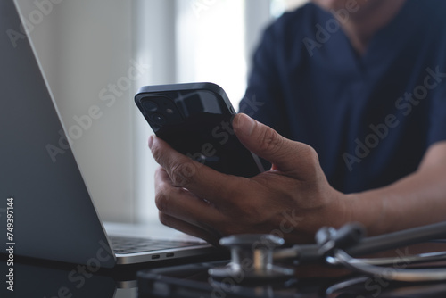 Doctor using mobile smart phone, working on laptop computer in medical workspace office with stethoscope on desk, close up, teleconference, telemedicine, medical research and technology concept photo