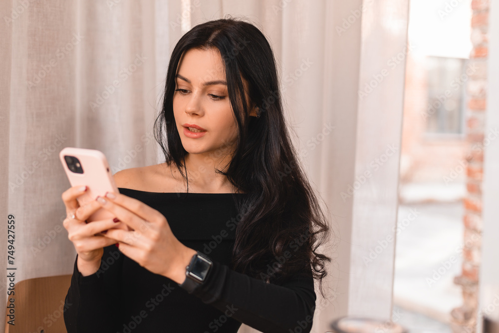 Portrait of calm teen female dressed casually holding mobile phone, typing messages, communicating with friends via social networks, using high Internet connection at cafe. Woman look flirting.