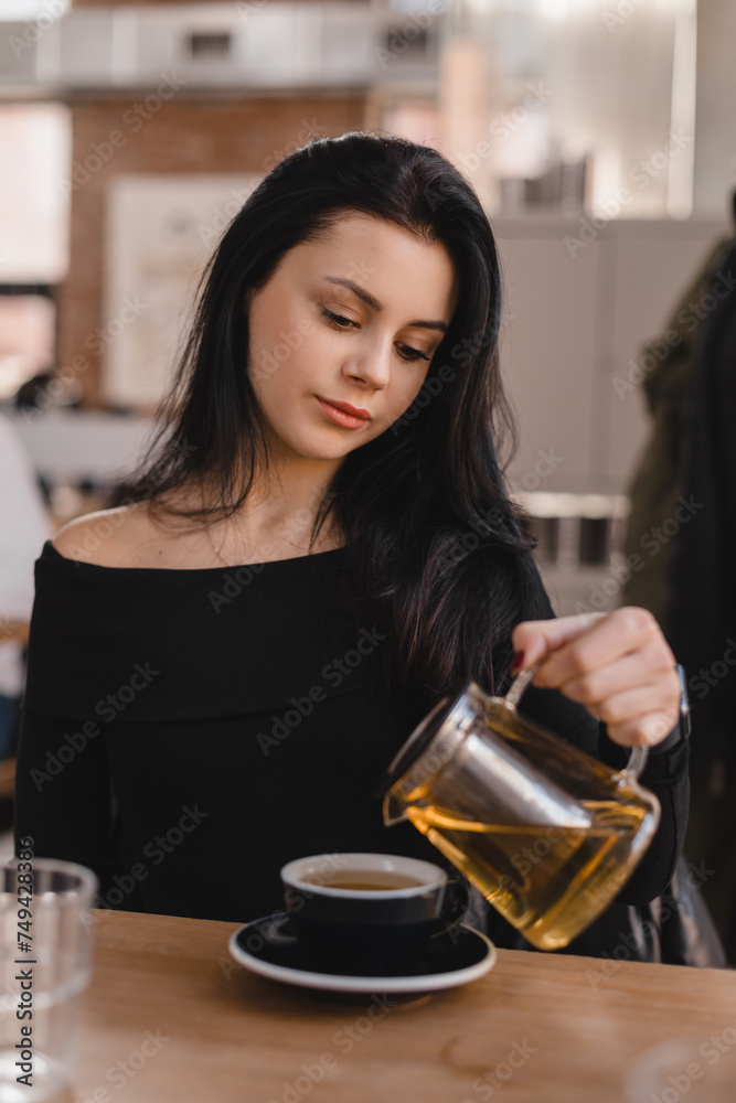 Brunette woman sitting at table drinking tea in restaurant. Teen woman wear black long sleeves top pouring green tea from glass teapot into cups in cafe. Happy girl relaxing and rest with friends. Brunette woman sitting at table drinking tea in restaurant. Teen woman wear black long sleeves top pouring green tea from glass teapot into cups in cafe. Happy girl relaxing and rest with friends.