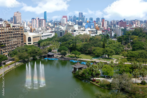 Aerial scenery of Downtown Taichung, a vibrant metropolis in central Taiwan, with modern skyscrapers in background & a rainbow fountain in a pond surrounded by green forests in a park under sunny sky
