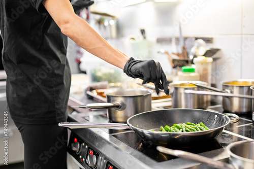 A chef in black attire sautés green vegetables in a bustling kitchen. Stainless steel pots and pans surround the stove. 