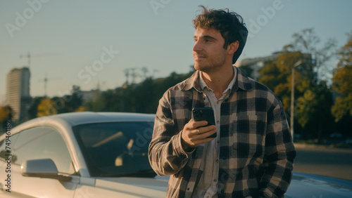 Successful Caucasian man stand near luxurious car in sunlight sunbeam outdoors happy male businessman hold smartphone browsing mobile phone internet buying new auto modern automobile dream in city