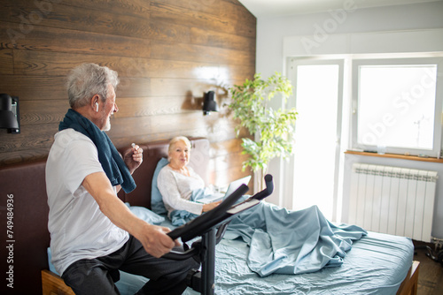 Happy senior couple in bedroom with woman using laptop and man exercising