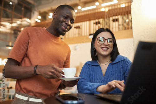 Waist up portrait of two smiling young people looking at laptop screen in coffee shop Black man and Middle Eastern woman