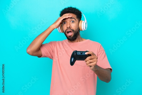 Young brazilian man playing with a video game controller isolated on blue background doing surprise gesture while looking to the side