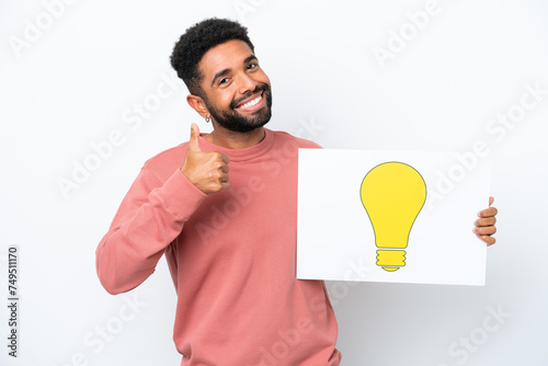 Young Brazilian man isolated on white background holding a placard with bulb icon with thumb up