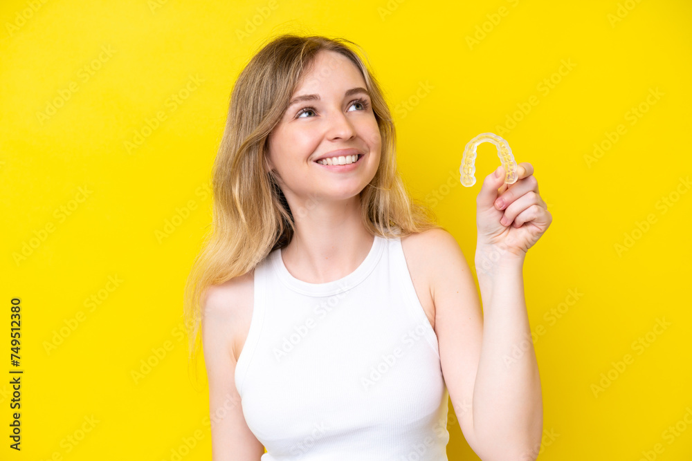 Blonde English young girl holding invisible braces isolated on yellow background looking up while smiling Blonde English young girl holding invisible braces isolated on yellow background looking up while smiling
