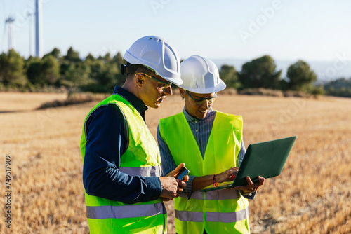 Engineers in protective work clothes and hard hats stand in a field of windmills.