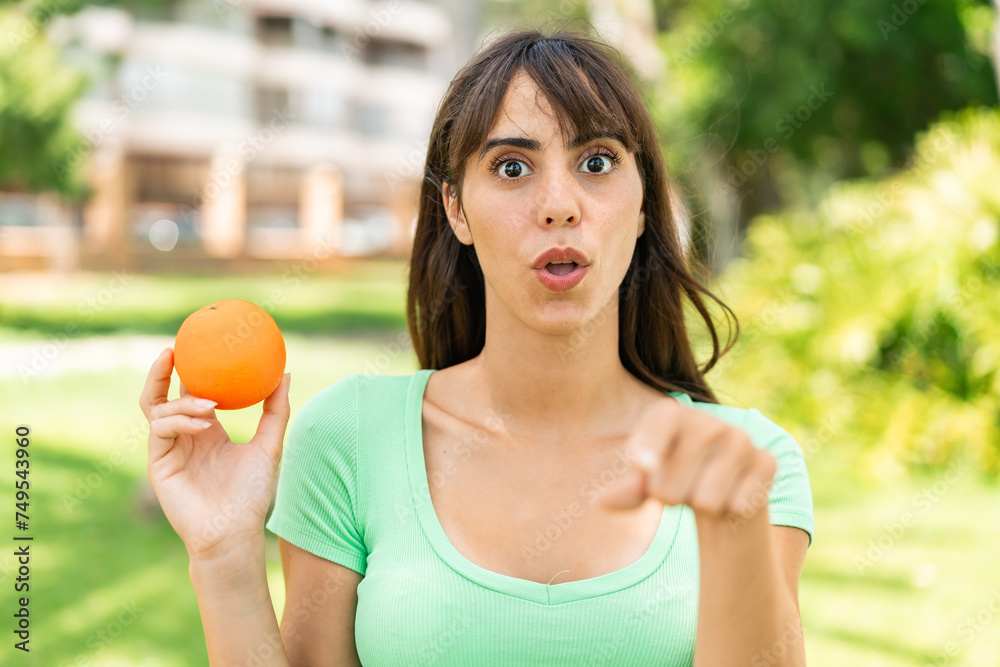 Young woman holding an orange at outdoors surprised and pointing front Young woman holding an orange at outdoors surprised and pointing front