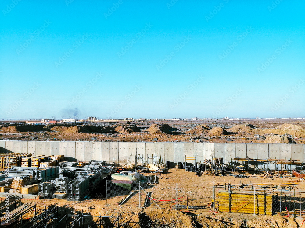 A construction site with a large wall in the background. The sky is clear and blue. Scene is calm and peaceful