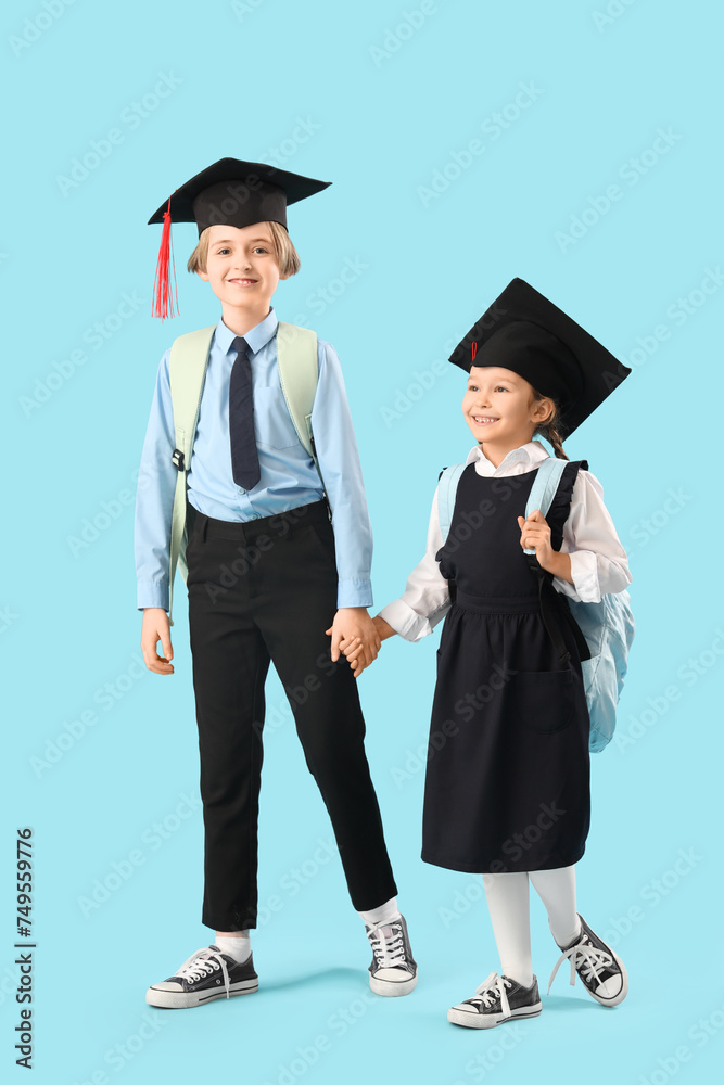 Little children in graduation hats with backpacks walking on blue background. End of school year Little children in graduation hats with backpacks walking on blue background. End of school year