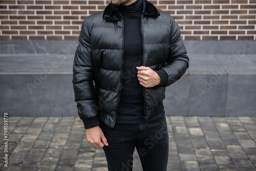 A man in a stylish leather jacket with a collar stands against the background of a brick wall. Fashionable branded luxury clothing for men