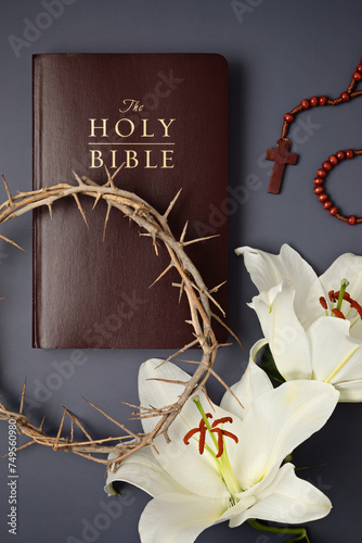 A symbolic Christian composition with Holy Bible lies beside a crown of thorns and a fresh white lily on a dark background as traditional Easter symbols.