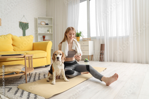 Sporty young woman with cute Beagle dog and mobile phone after doing yoga on mat at home