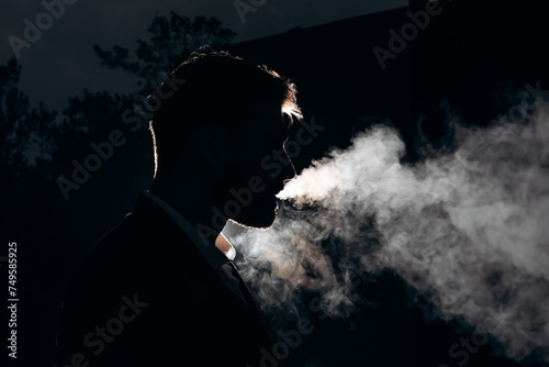 Dark photo of a silhouette of a man blowing smoke against the light. Black background and white smoke. Night portrait of a smoking man photo
