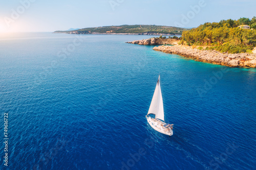 Aerial view of sailboat on blue sea at sunset in summer. Travel in Sardinia, Italy. Tropical seascape with sailing boat, sea bay, mountain, green trees, ocean, sky. Top drone view yacht. Yachting