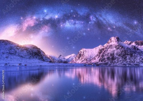 Milky Way arch, sea coast and snow covered mountains in winter at night. Lofoten Islands, Norway. Arctic landscape with starry purple sky, arched milky way reflected in water, snowy rocks. Space