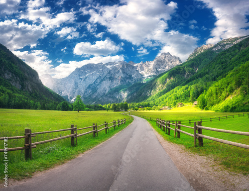Rural road in alpine mountains, wooden fence, green meadows, trees in summer in Logar valley, Slovenia. Country road. Colorful landscape with road, rocks, field, grass, blue sky with clouds at sunset 