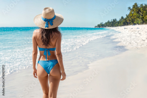A woman in a blue bikini and straw hat stands on a beach.. Young sexy woman at tropical beach. © vachom