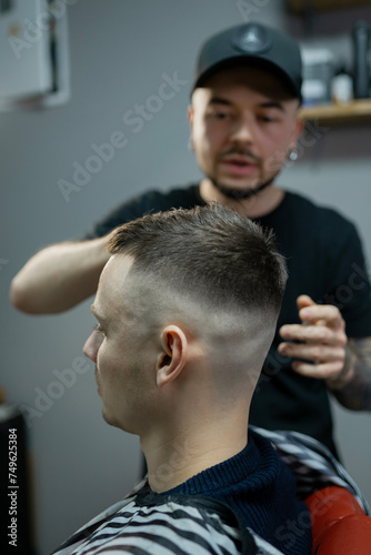 A barber man with a tattoo on his arm makes a haircut to a brunette guy in a barbershop, cuts his hair with a clipper close-up