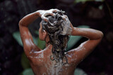 A naked woman takes a shower in an outdoor shower with tropical plants. A shampoo advertisement in dark colors.