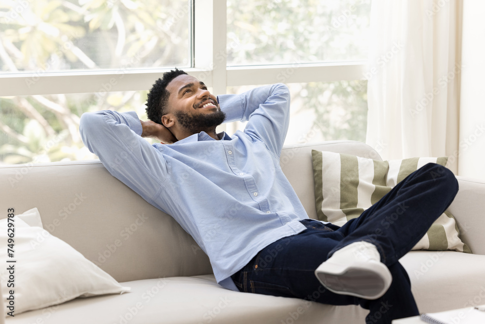 Happy carefree young African man relaxing on cozy home couch, stretching body on soft back with open elbows, breathing fresh cool air, enjoying success, comfort, looking away, daydreaming