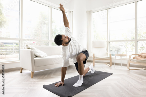 Fototapeta Naklejka Na Ścianę i Meble -  Serious young Black man doing morning exercises at home, keeping twisting yoga asana on mat, caring for wellness, fit, wellbeing, healthy active lifestyle, training body in apartment
