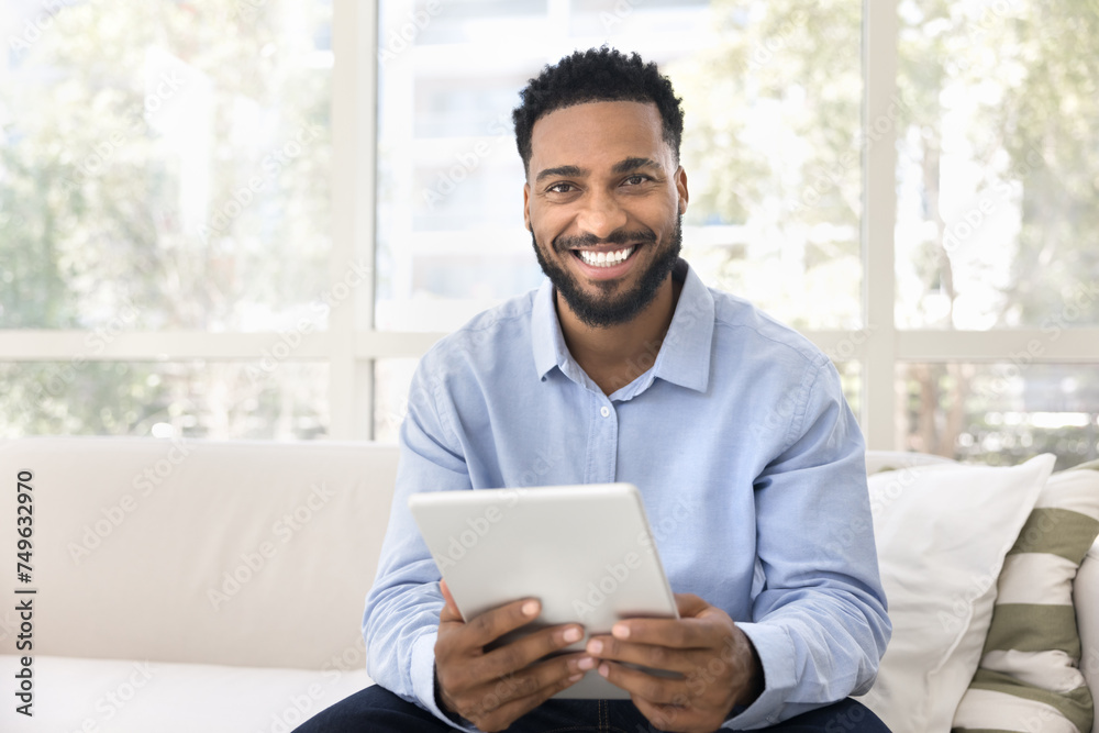Happy handsome young Black man holding tablet, looking at camera with toothy smile, sitting on white couch, using online application, financial service, enjoying communication, posing for portrait Happy handsome young Black man holding tablet, looking at camera with toothy smile, sitting on white couch, using online application, financial service, enjoying communication, posing for portrait