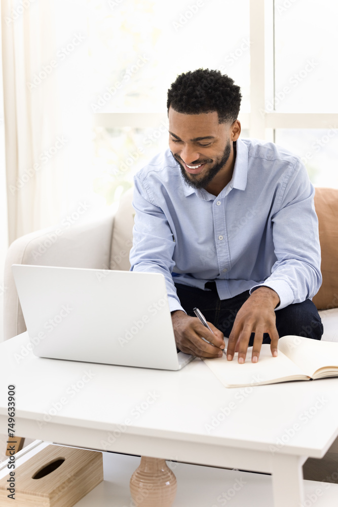 Cheerful young Black student guy watching online training lecture on laptop, writing notes, speaking on educational Internet conference meeting, using notebook. Vertical shot Cheerful young Black student guy watching online training lecture on laptop, writing notes, speaking on educational Internet conference meeting, using notebook. Vertical shot