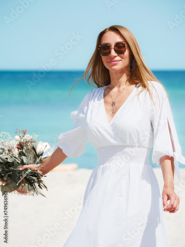 Obraz na płótnie Joyful Young Woman Holding a Bouquet of Flowers on a Beach with Blue Sky Background w salonie