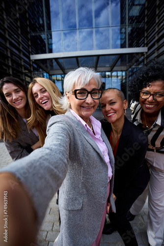 Vertical group of diverse business formal women taking selfie with phone looking smiling at frontal camera. Happy cheerful photo of adult female people co-workers outdoor. Gray mature lady in center photo