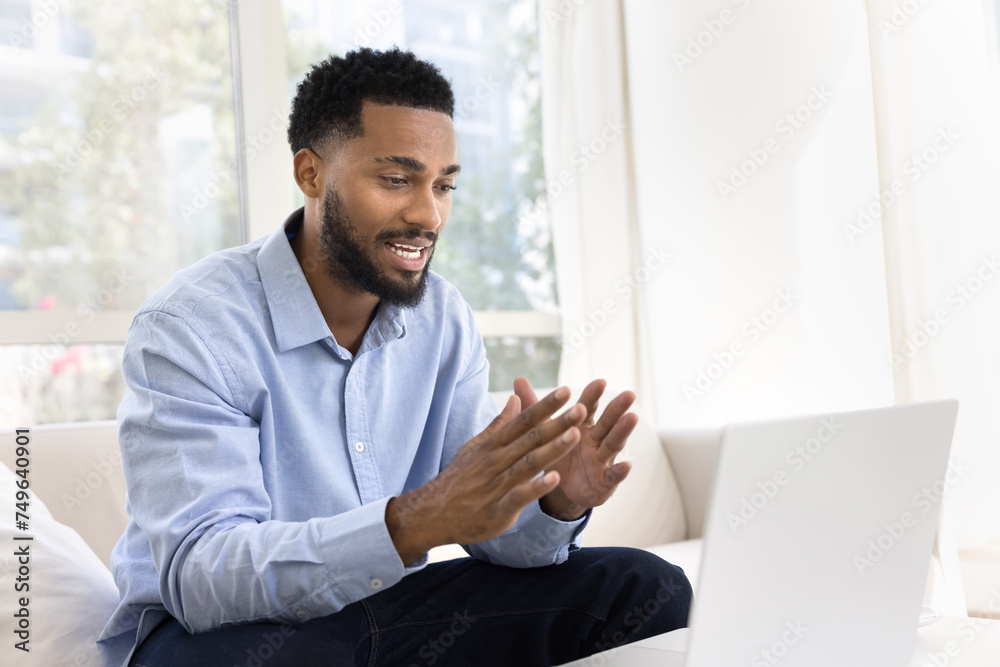 Fototapeta premium Positive handsome young African business man talking on work video call on laptop, speaking at computer, moving hands, convincing client on online conference meeting