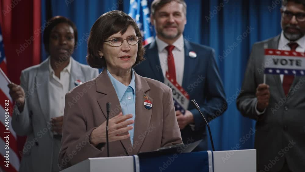 Medium shot of mature Caucasian female presidential candidate from Republican or Democratic party standing at podium, presenting political manifest at primary elections, diverse team in background Medium shot of mature Caucasian female presidential candidate from Republican or Democratic party standing at podium, presenting political manifest at primary elections, diverse team in background