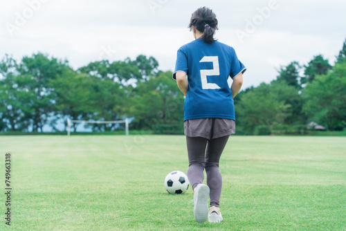 グランドでサッカーボールをドリブルするユニフォームを着た女性の後ろ姿
