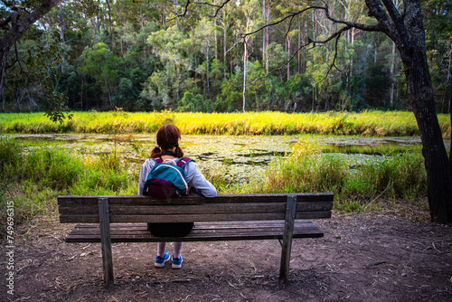 pretty girl with a backpack resing on a bench enjoys the view of enoggera reservoir in brisbane, queensland, australia; warm sunset in a park	 photo