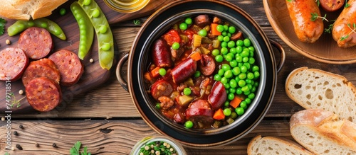 A variety of food items are displayed on a wooden table, including a bowl of sausage vegetable stew, green peas, fried sausage slices, and wheat bread. The view is from above.