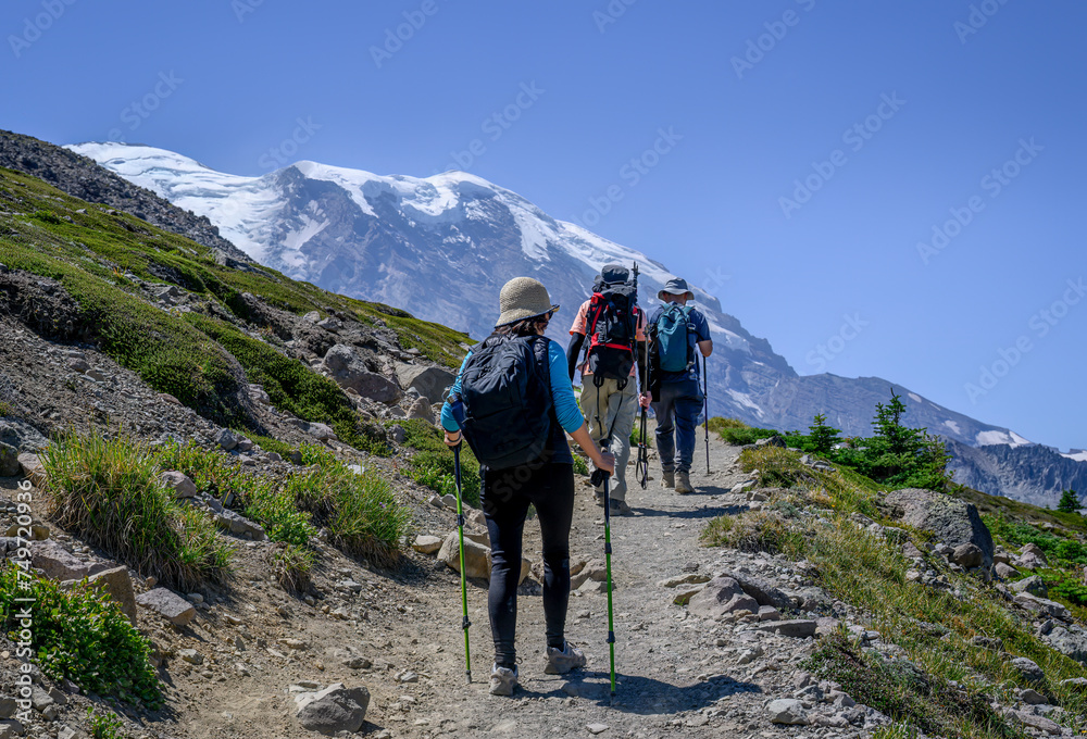 Three people hiking Sunrise Trail in summer. Snow-capped Mount Rainier looming large in front. Washington State.