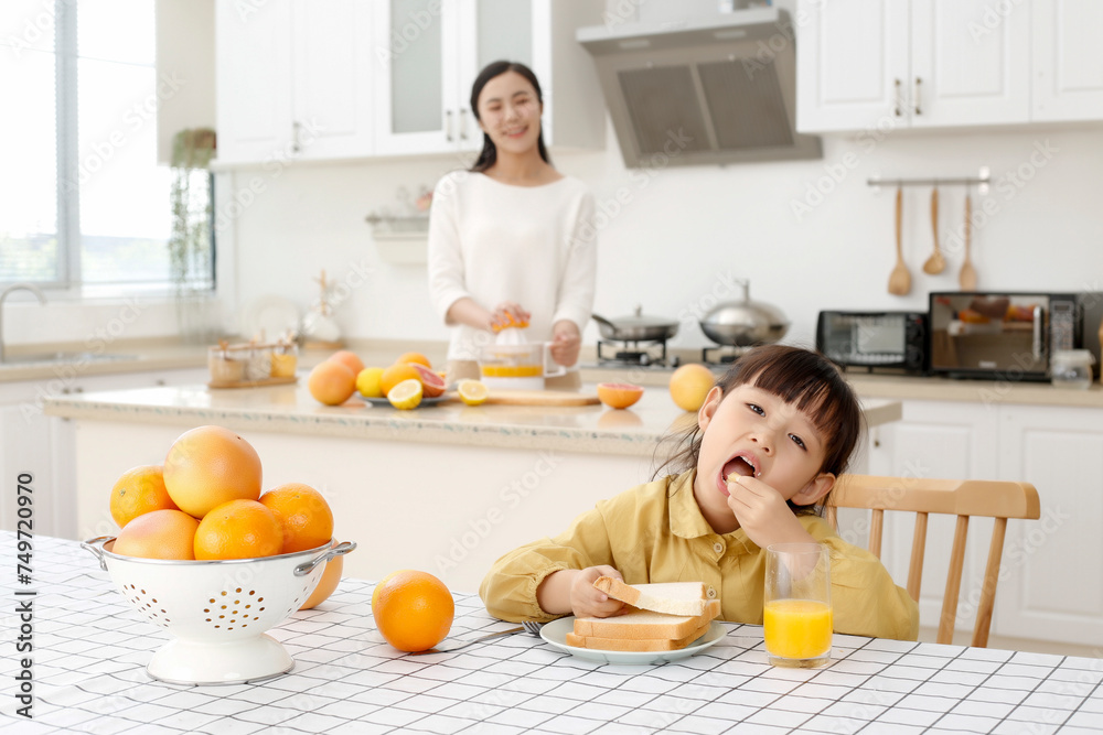 Asian woman making orange juice in kitchen, girl drinking juice and eating bread