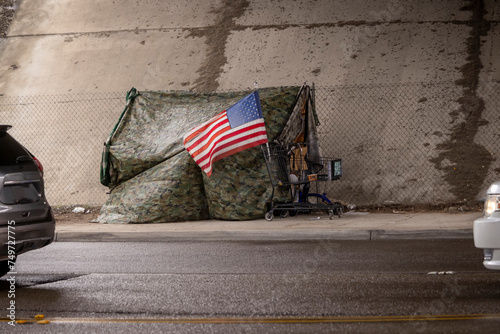 An American flag at a homeless tent made of camouflage tarp at a road underpass with a car visible photo