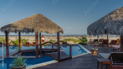 A recreation area by the ocean. There are sun loungers and straw umbrellas on the boardwalk by the swimming pool. A man stands and looks at the sea. A row of sunbeds under a parasols on a sandy beach. © Вера 