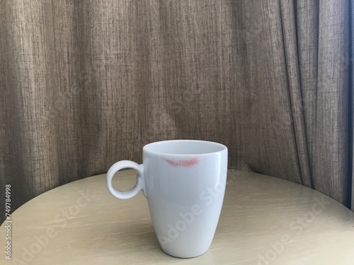 white mug of coffee with red lipstick mark put on brown table and gray curtain on background with morning sunlight shining from the right side of frame