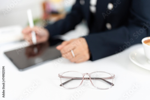 Close-up of glasses on a desk and a business woman in a long-sleeved suit wearing a ring. Have coffee mug, use laptop and pen to do business work in cafe of stock exchange company