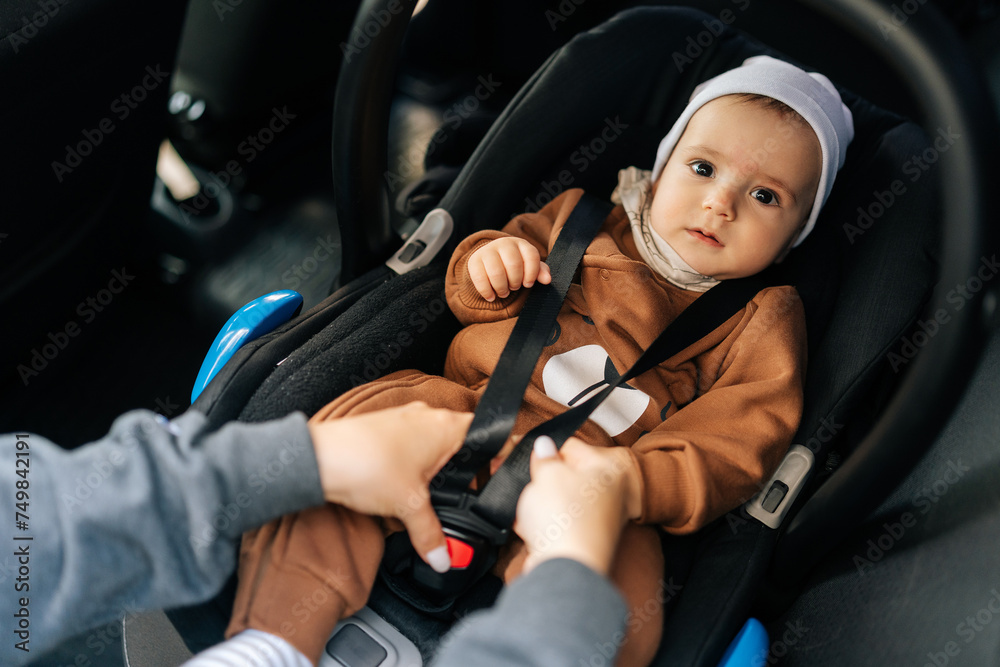 Portrait of adorable infant baby boy which unrecognizable young mother fastening safety belt in car seat. Cute toddler with security belt in vehicle. Concept of safety lifestyle, driving and travel.