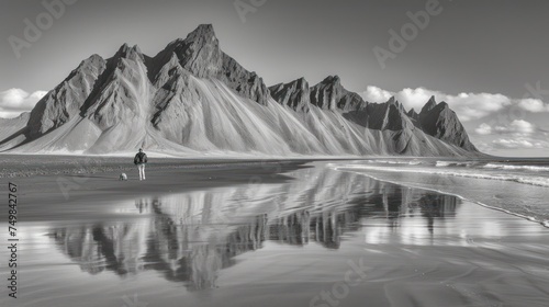 a black and white photo of a person standing in front of a mountain with a reflection of them in the water. photo