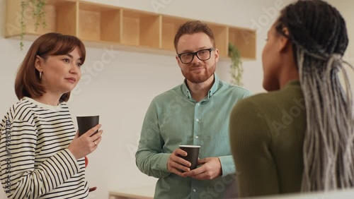 Three friendly multiethnic coworkers standing in office, drinking coffee and talking cheerfully in workspace during break. Male and female colleagues having conversation at modern open space. photo
