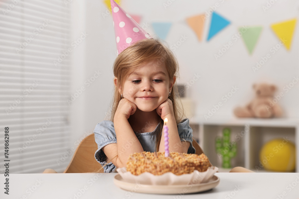 Cute girl in party hat with birthday cake at table indoors Cute girl in party hat with birthday cake at table indoors
