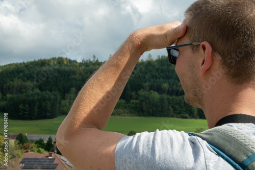 Side view of a young man looking far, looking at the horizon with his hand on his forehead. Beautiful nature with forest and clouds. Travel and forecast concept. photo