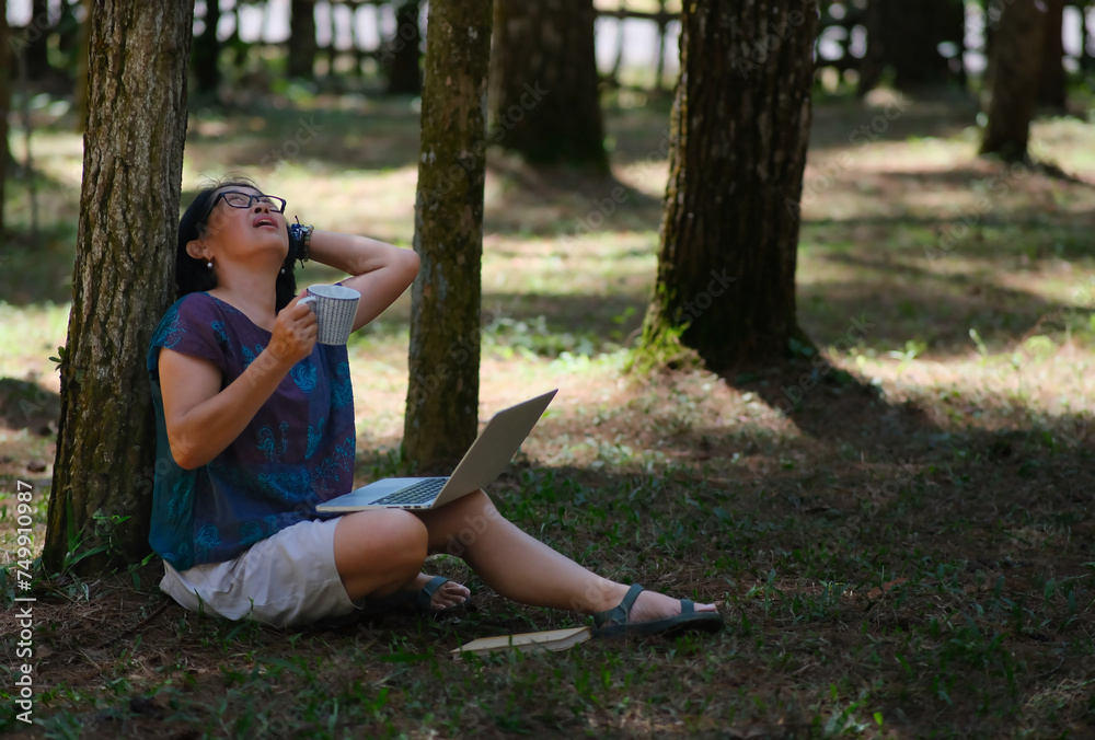 Woman sitting on the grass curiously looking up a tree above her, while working on her computer and enjoying tea Woman sitting on the grass curiously looking up a tree above her, while working on her computer and enjoying tea