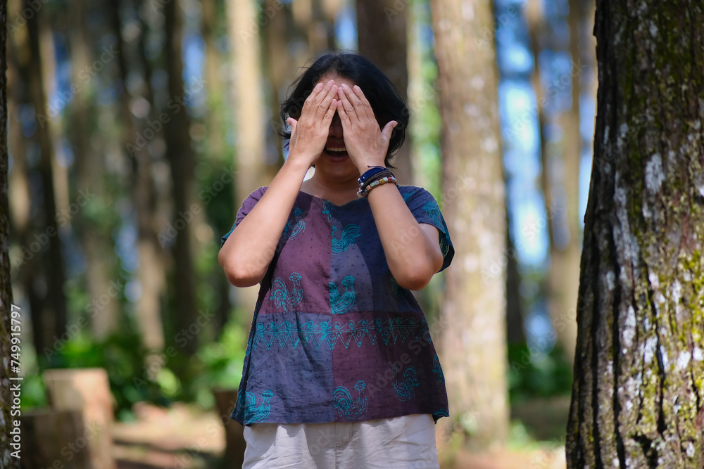 Woman standing alone in the park, covering her face with both hands, playing hide and seek Woman standing alone in the park, covering her face with both hands, playing hide and seek