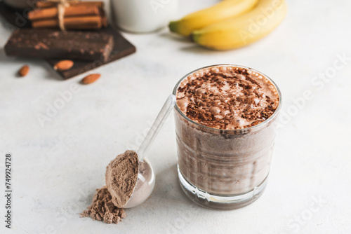 Glass with chocolate protein drink, milkshake smoothie on white table with bananas, protein powder in measuring spoon, protein bar, almond nuts and cinnamon sticks